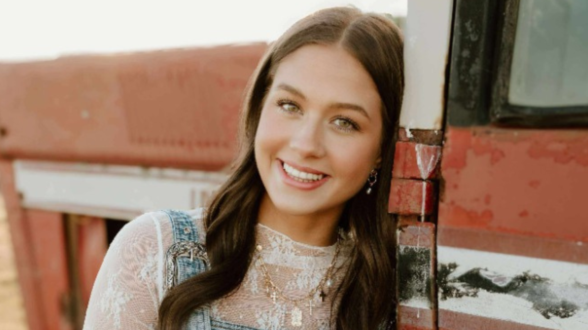 A young woman with long brown hair, wearing denim overalls over a lace top, smiles while leaning against a weathered red truck.